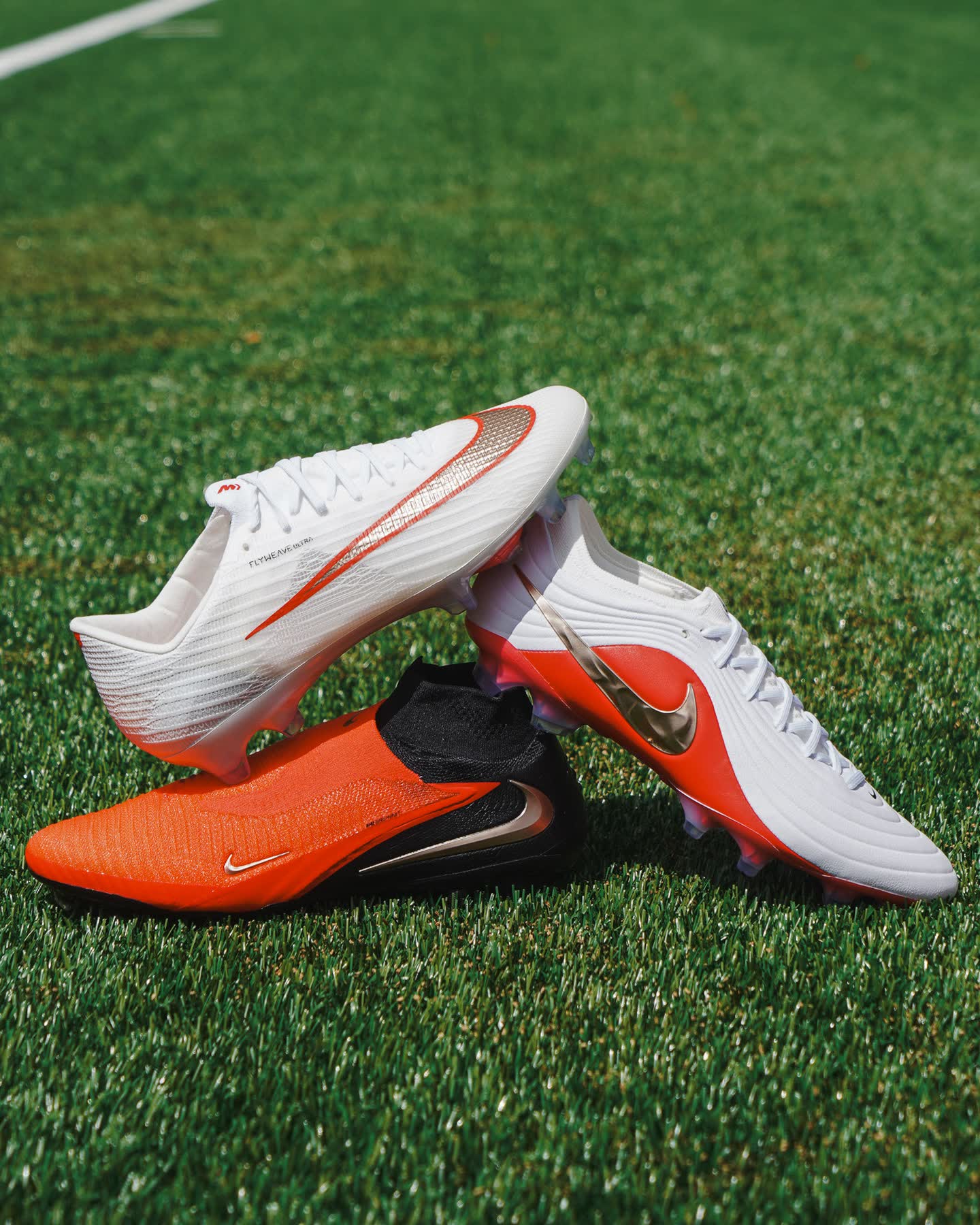 Помаранчевий Nike Phantom 6 High з чорною в’язаною манжетою, на ньому дві білі низькі бутси Nike, на траві